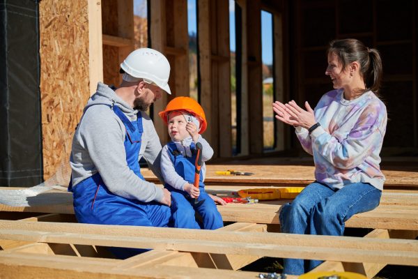 Father, mother and son building wooden frame house. Toddler boy helping his daddy, while woman looking for them on construction site. Guys wearing helmet and blue overalls. Carpentry, family concept.